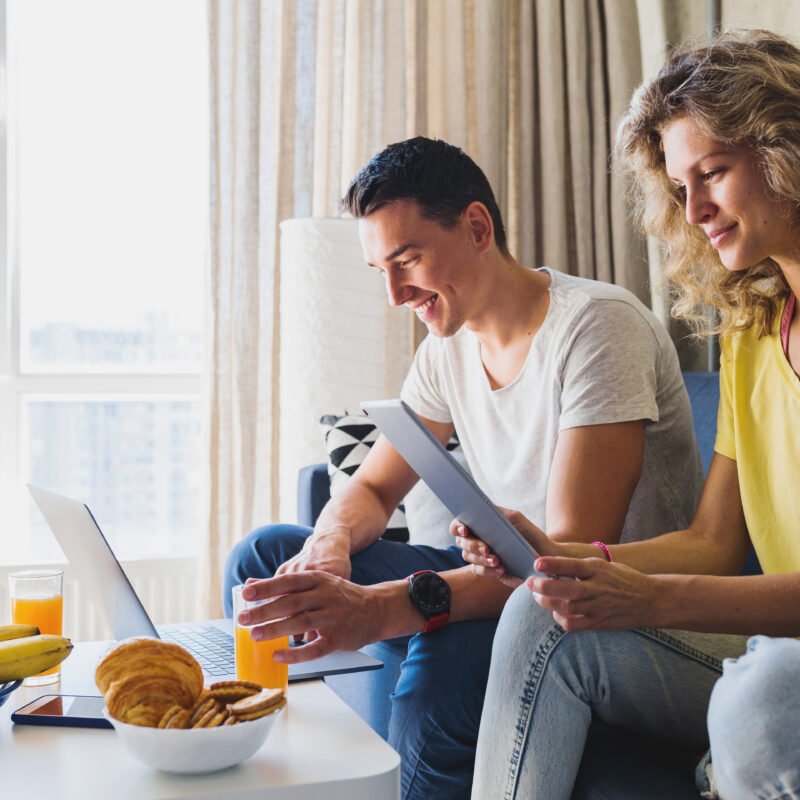 Imagen de Young couple sitting on sofa at home working online on laptop and tablet, using internet, smiling, happy together, having fun, man and woman in love, family leisure time, having breakfast in morning