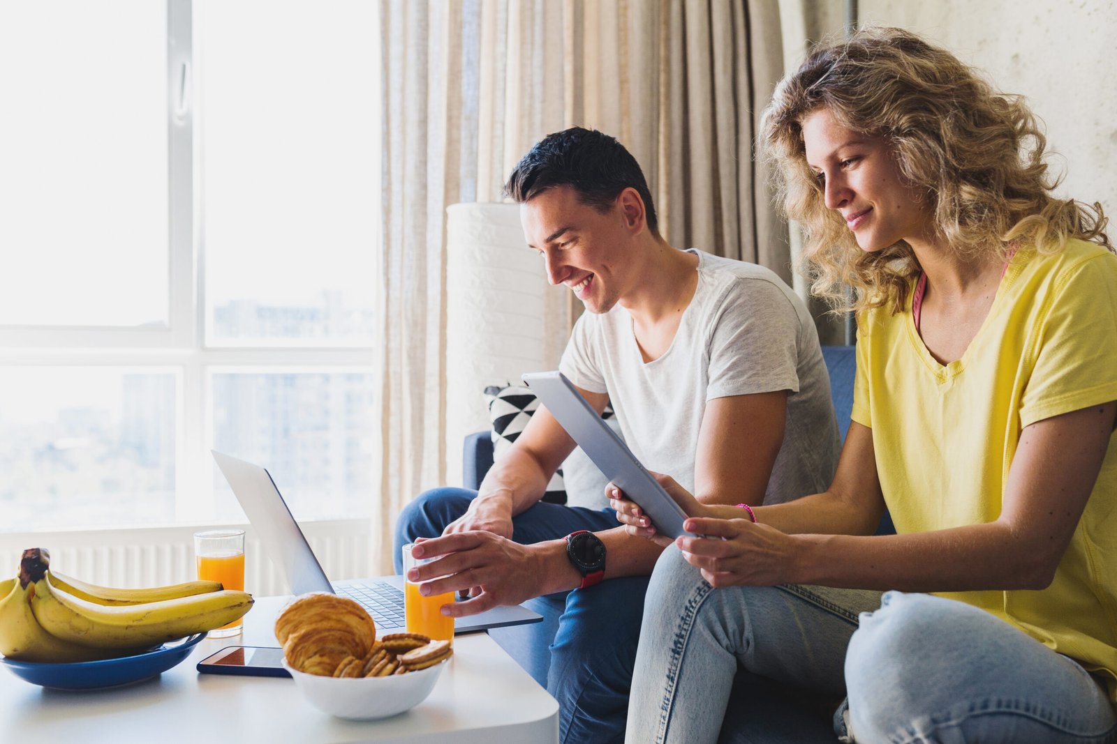 Imagen de Young couple sitting on sofa at home working online on laptop and tablet, using internet, smiling, happy together, having fun, man and woman in love, family leisure time, having breakfast in morning